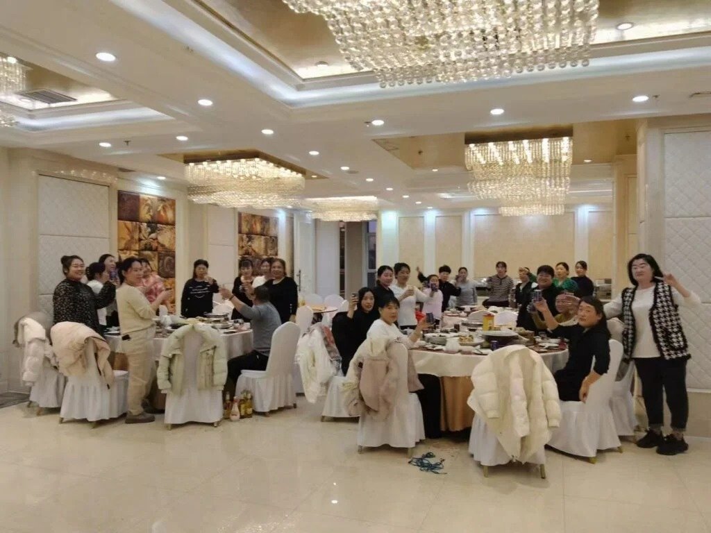 Group of people celebrating at a banquet in a luxury hall with chandeliers, white tablecloths, and winter coats draped over chairs.