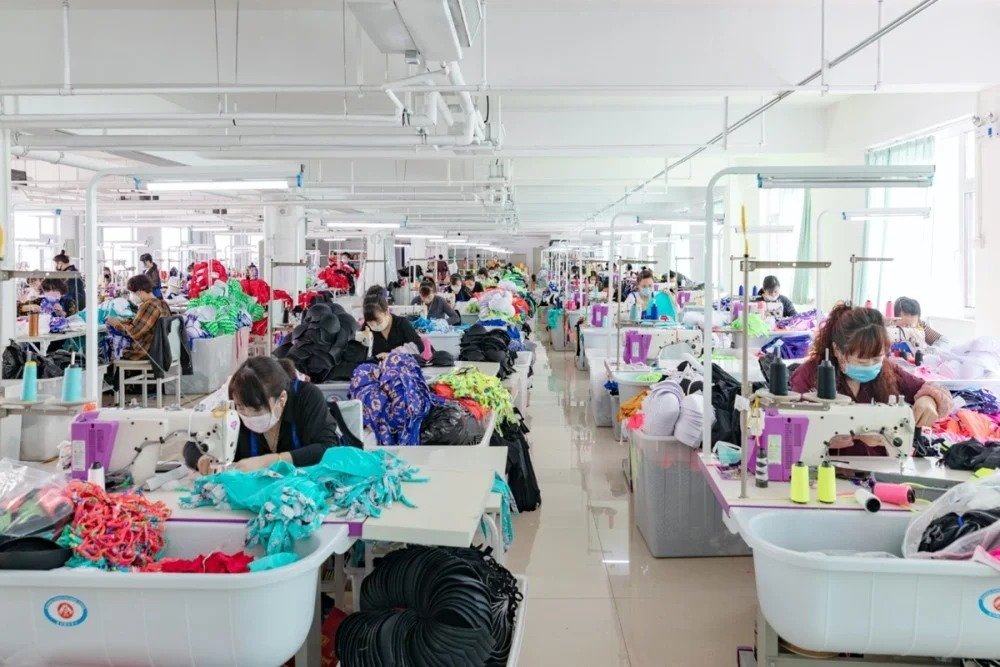 Workers in masks operate sewing machines producing colorful swimwear and activewear in a bright, organized factory production line with fabric piles and spools of thread.