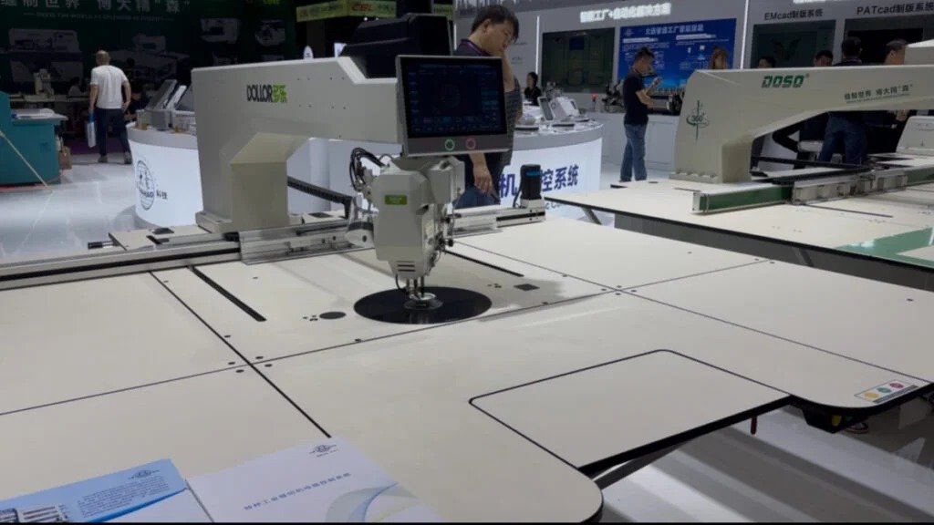Industrial embroidery machine in action at trade show, white DOLCE and DOSEO models for swimwear manufacturer production, showcasing automated sewing system with digital control panel.
