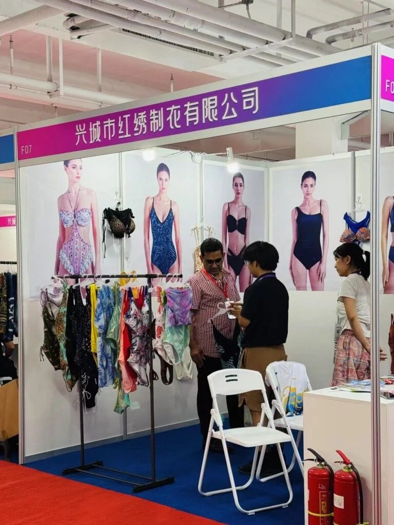 Hongxiu Clothing booth at trade show displaying colorful swimwear including floral and solid one-pieces, with staff discussing production details near rack of bikinis and trunks.
