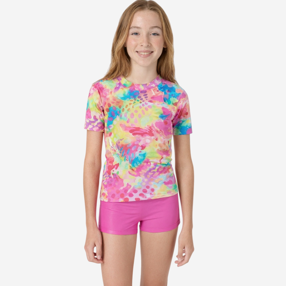 Young girl wearing vibrant tie-dye short-sleeve rash guard in pink, blue, yellow, and green with polka dot accents, paired with shiny fuchsia swim shorts.