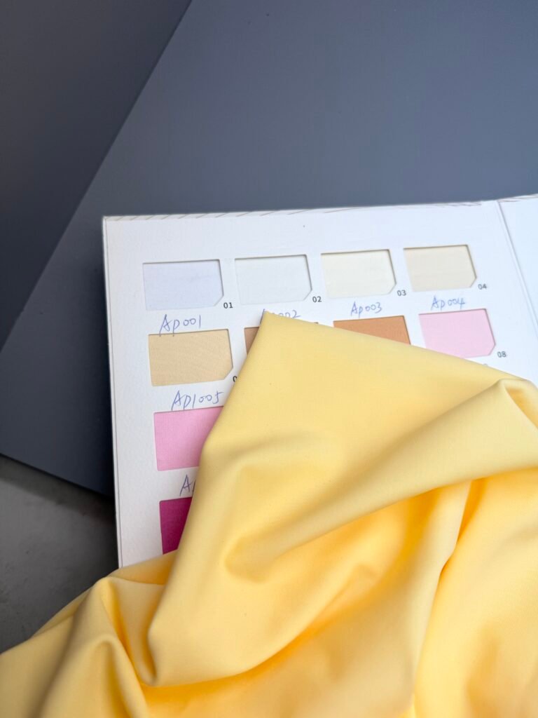 Yellow stretch swimwear fabric sample (Ap1005) displayed over color swatch book with labeled shades for B2B swimwear manufacturer production.