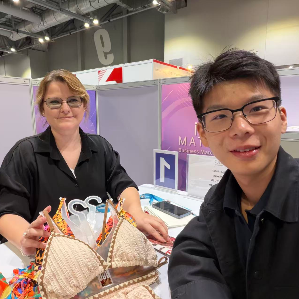 Two people at a trade show booth displaying custom swimwear bikini tops on hangers.