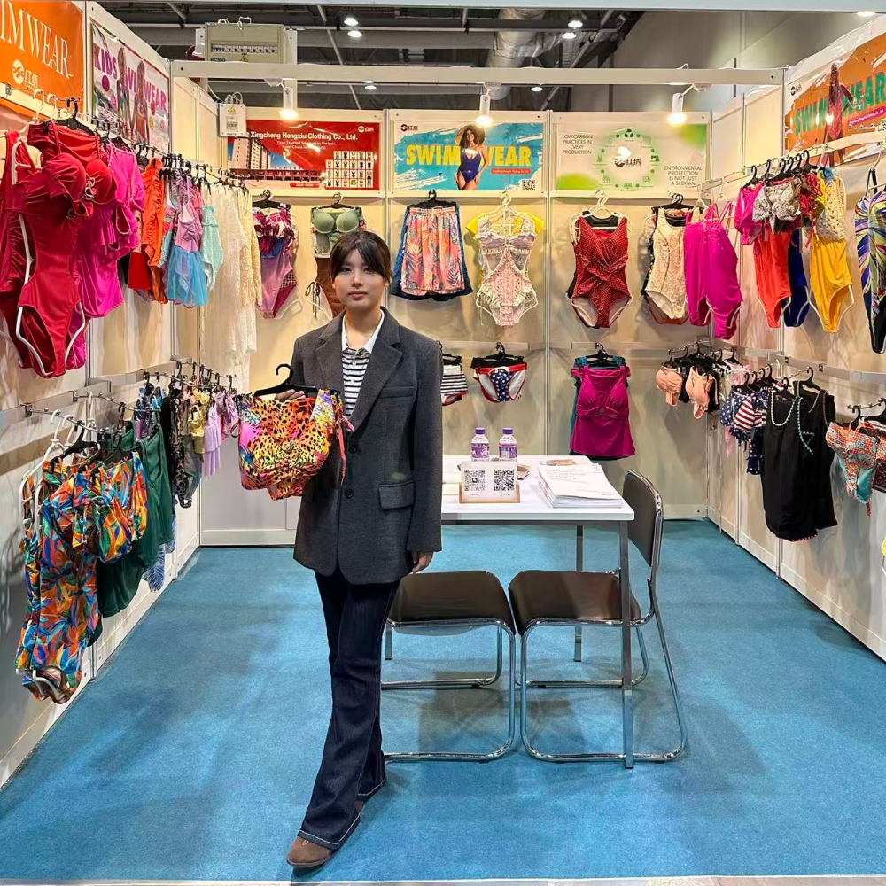 A woman holds a colorful swimsuit at a swimwear manufacturer trade show booth displaying various garments.