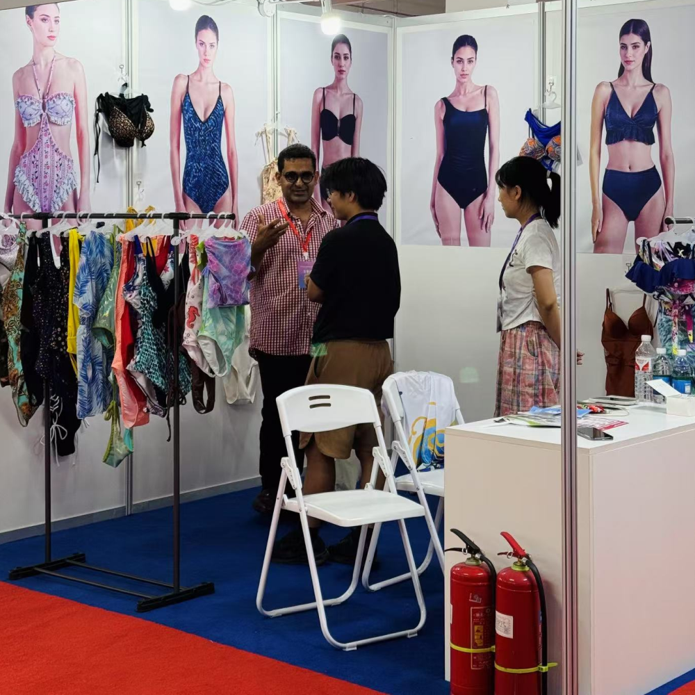People discussing swimwear samples at a trade show booth with racks of garments and model posters.