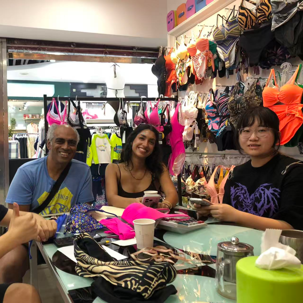 Three people sitting at a table in a swimwear showroom surrounded by colorful bikinis on display racks.