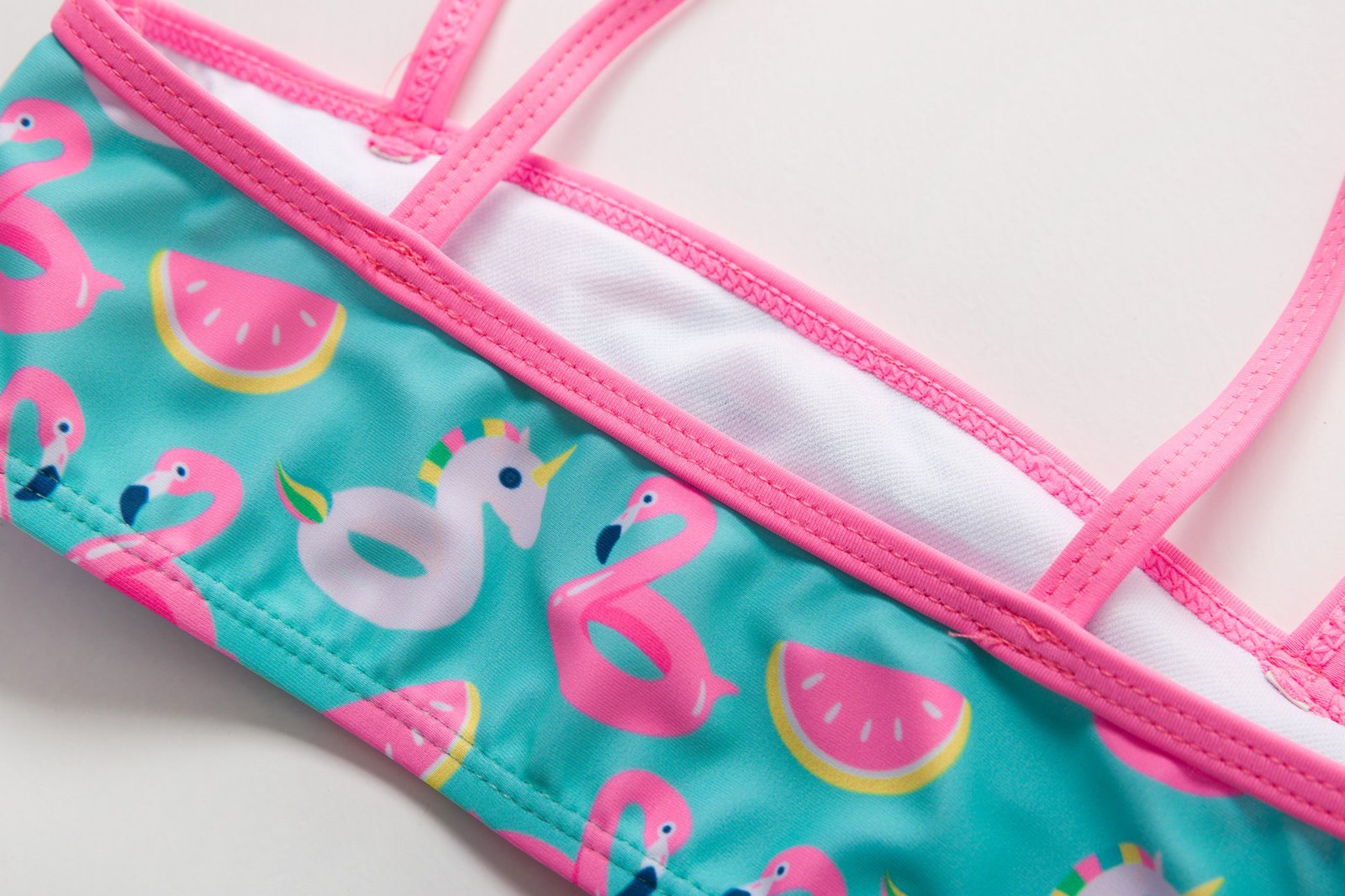Close-up of a teal girls' bandeau bikini top featuring flamingo, watermelon, and unicorn prints with pink trim and