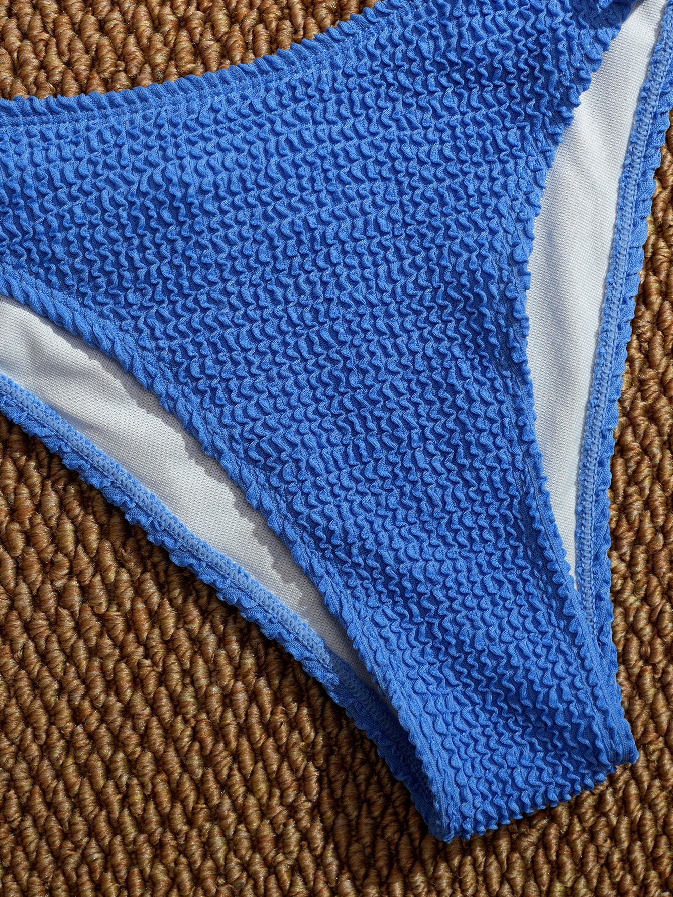 Blue textured triangle bikini bottom with white lining on woven mat.