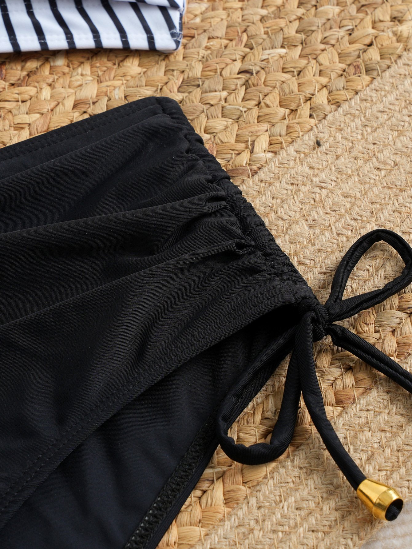 Close-up of black high-waist bikini bottoms with side tie straps and gold aglets on woven mat.