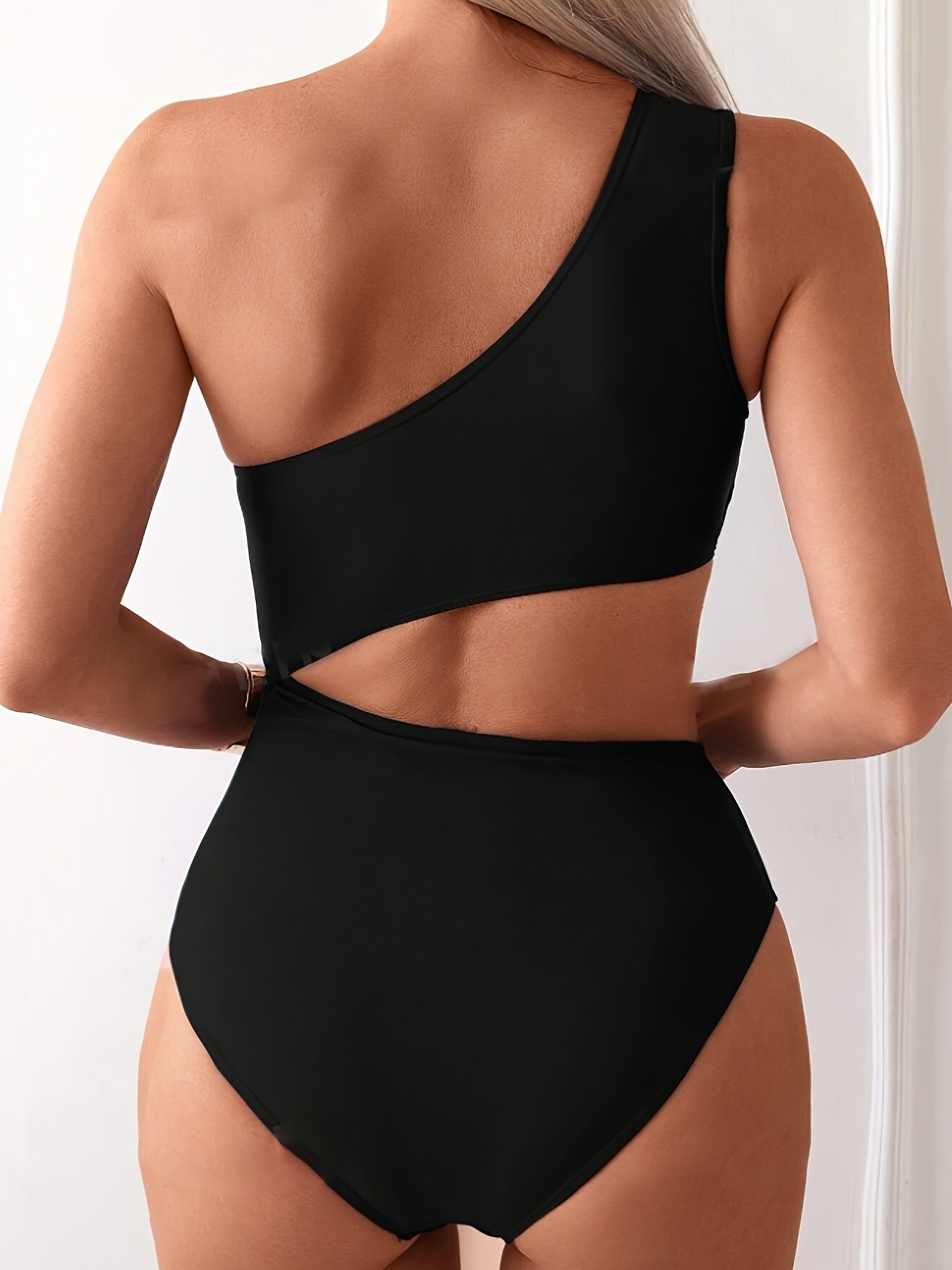 Back view of a black one-shoulder monokini featuring a diagonal midriff cutout and high-cut leg design.
